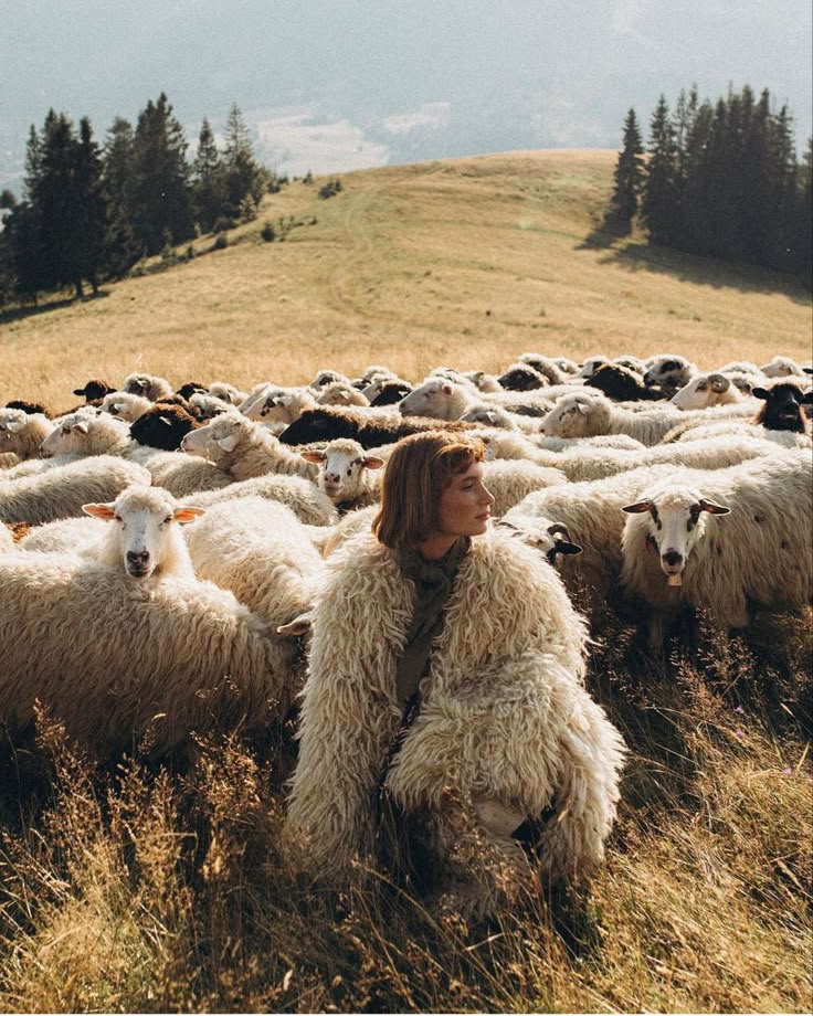 A woman in a soft fleece coat sitting among a herd of sheep on a hillside